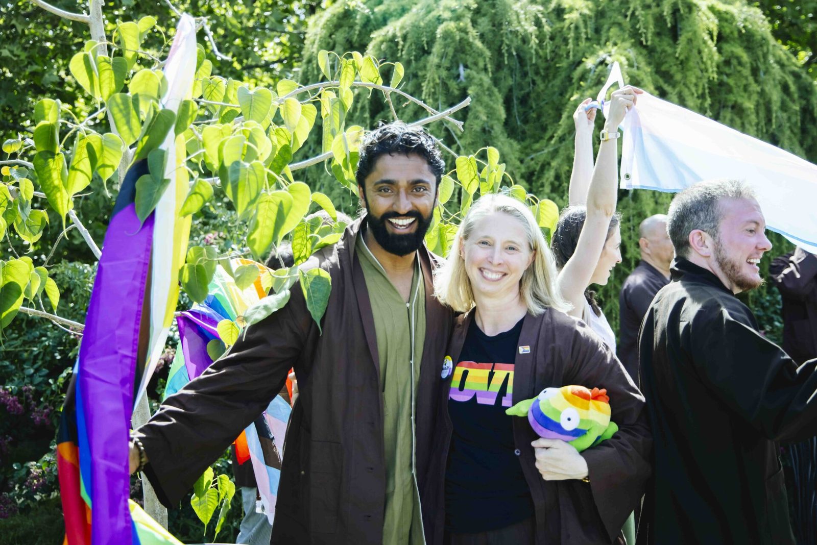 Two friends standing arm in arm outdoors, smiling, with rainbow flags and streamers hanging in the trees behind them.