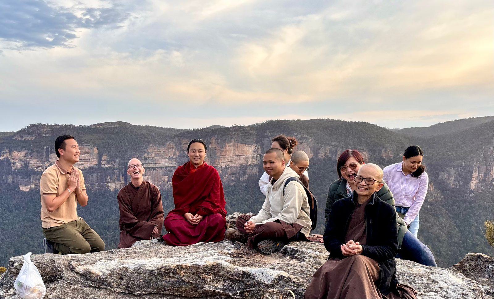 Nine friends, several of them in monastic robes, sitting and kneeling along a sandstone ledge at sunset, laughing together, with a forested valley and cliffs behind them.