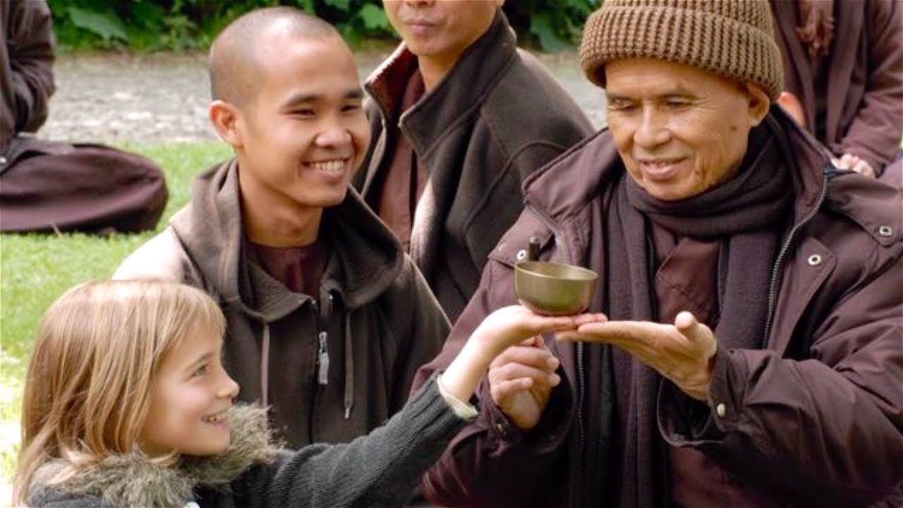 Thich Nhat Hanh, in a knitted brown hat and winter coat, holding a small bell on his open palm while a young girl reaches up to touch it. A smiling young monastic sits beside them and two more are behind, on grass under trees.
